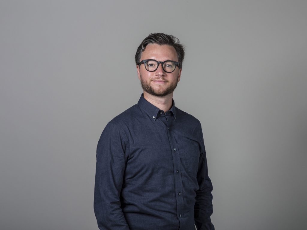 A headshot of Max Oglesbee against a grey background. Max has short, neat hair, dark rimmed glasses, and a dark blue button down shirt.